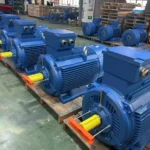 Several large blue industrial electric motors lined up on wooden pallets in a factory setting.