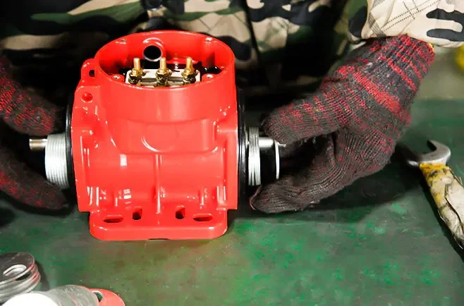 Componente industrial vermelho com múltiplas conexões sendo manuseado por um trabalhador usando camuflagem e luvas em uma superfície plana.