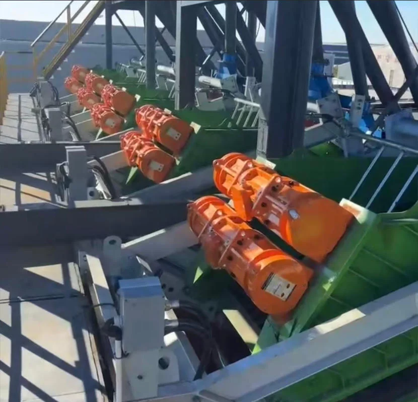 Fileira de telas vibratórias laranja em uma instalação industrial verde. Os dispositivos estão alinhados paralelamente, situados numa plataforma exterior.