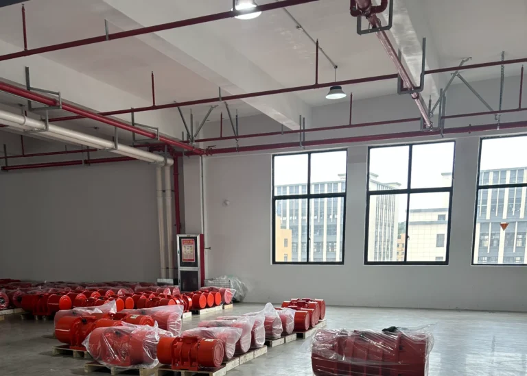 Interior do armazém com equipamento industrial vermelho embrulhado em plástico, arrumado em paletes de madeira sob grandes janelas.