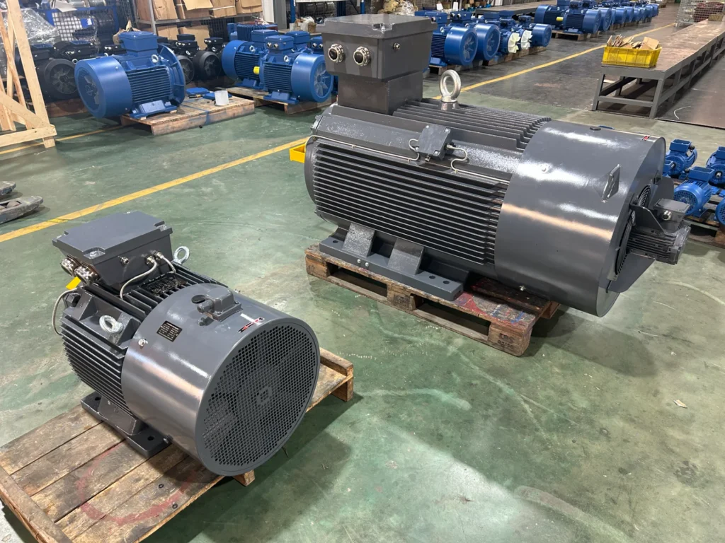 In einer Lagerhalle mit Betonboden sind mehrere industrielle Elektromotoren auf Paletten platziert. Die Motoren variieren in der Größe und sind dunkelgrau und blau lackiert.
