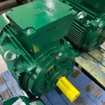 Green industrial electric motor with a yellow shaft resting on a blue pallet in a workshop setting.