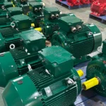 Rows of green and red industrial electric motors organized on pallets in a warehouse setting with a worker in the background.